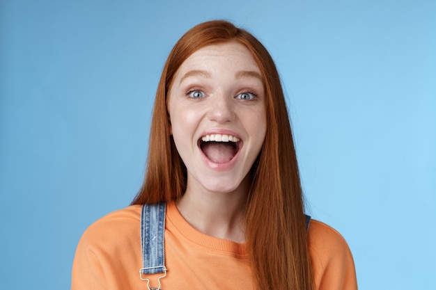 Fille de gingembre sortante animée charismatique amusée ravie de s'amuser des amis criant dire un sourire blanc parfait montrant une atmosphère joyeuse amicale debout excité fond bleu positif