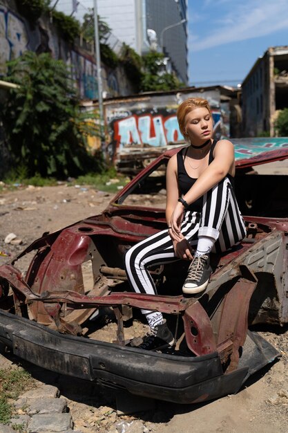 Fille glamour en pantalon et cheveux courts et maquillage posant près d'une vieille voiture cassée