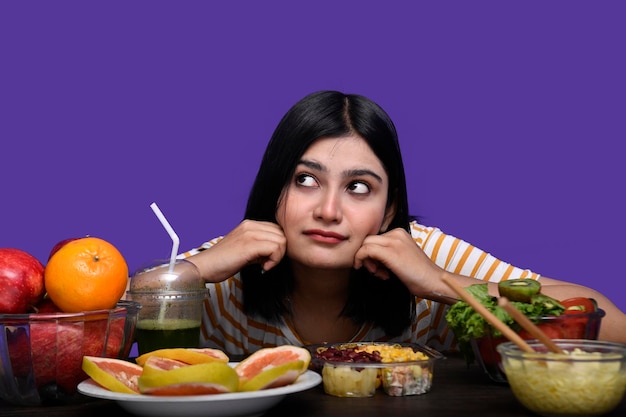 Fille gourmande assise à la table de fruits souriant les mains sur la table en levant le modèle pakistanais indien