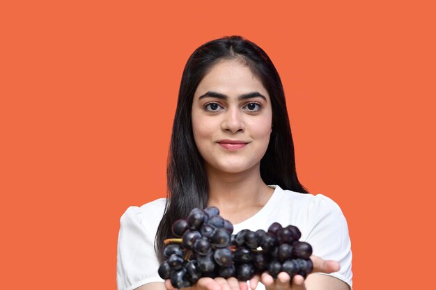 Fille gourmande portant un t-shirt blanc tenant des raisins et regardant la caméra modèle pakistanais indien