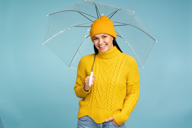 Fille gracieuse portant un élégant pull d'automne debout sous un parasol transparent et souriant. Portrait en studio d'un modèle féminin caucasien posant avec un parapluie sur fond bleu
