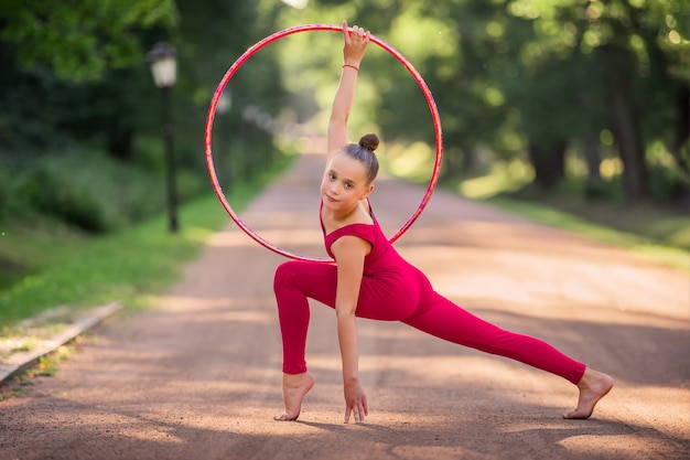 Une fille gymnaste dans un commando rouge fait un exercice avec un cerceau dans le parc un soir d'été