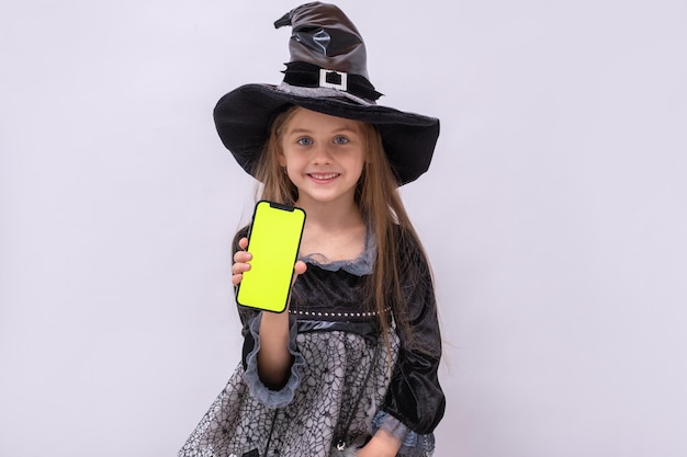 Fille d'Halloween sur fond gris Enfant en costume de sorcière Bannière d'Halloween heureuse avec espace de copie La fille tient un smartphone avec un écran vert Maquette Boutique Internet Achats en ligne avec ventes