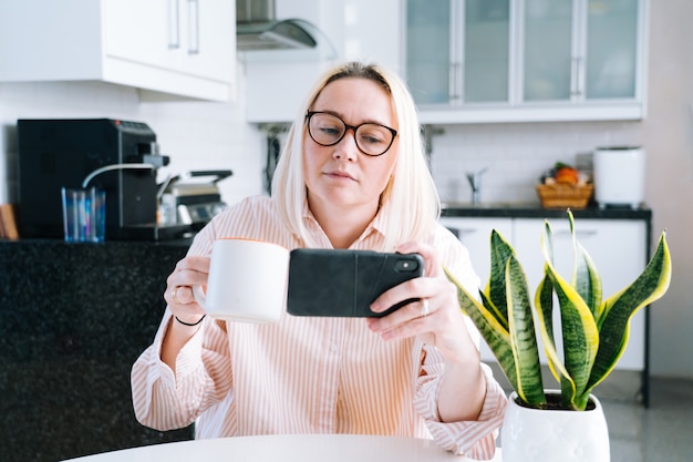 Fille Heureuse, Assis à La Cuisine à Domicile Et Tenant Un Appel Vidéo. Jeune Femme à L'aide De Smartphone Pour Appel Vidéo Avec Un Ami Ou En Famille. Webinaire D'enregistrement De Vlogger. Femme, Regarder, Appareil Photo, Et, Onduler, Salutation, Mains