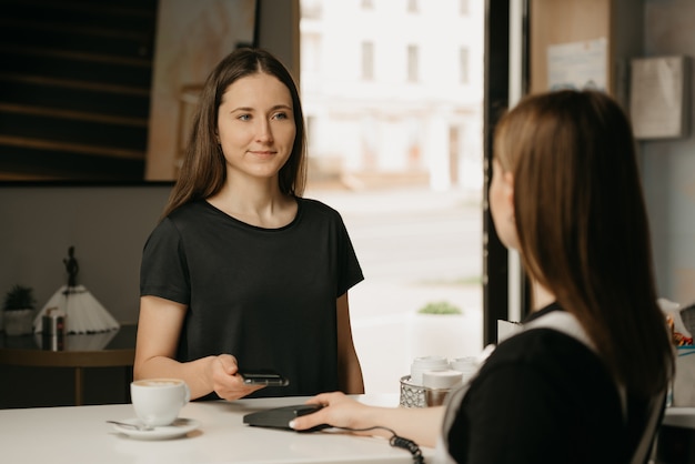 Une fille heureuse aux cheveux longs payant son café avec un smartphone par la technologie NFC sans contact dans un café. Une brune barista offre à un client un terminal pour le paiement.