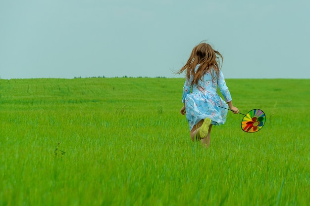 Une fille heureuse aux cheveux longs traverse un champ vert avec un moulin à vent dans les mains. Concept d'été et d'enfance heureuse.