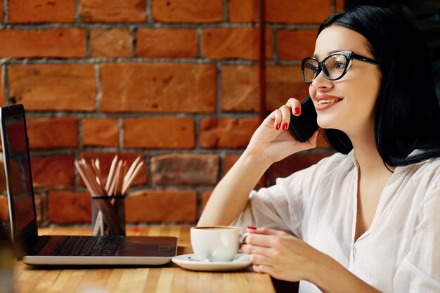 Fille heureuse aux cheveux noirs portant des lunettes assis dans un café avec ordinateur portable, téléphone portable et tasse de café, concept indépendant, portrait, espace copie, vêtu d'une chemise blanche.