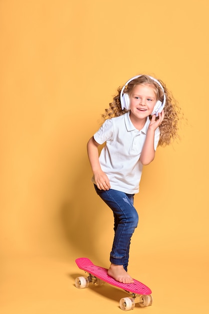 Fille heureuse avec des cheveux bouclés et des écouteurs s'amusant avec penny board