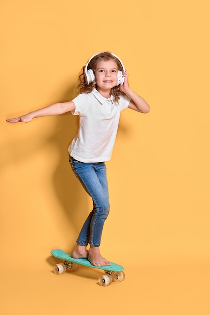 Fille heureuse avec des cheveux bouclés et des écouteurs s'amusant avec penny board