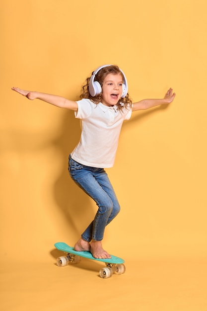 Fille heureuse avec des cheveux bouclés et des écouteurs s'amusant avec penny board