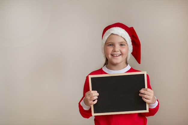 Fille heureuse en costume de père Noël tenant un bureau noir sur un mur gris clair