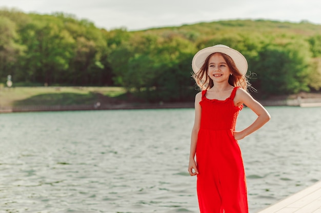 Fille heureuse dans une combinaison rouge et un chapeau blanc au bord de l'eau Plaisir de la liberté de la nature