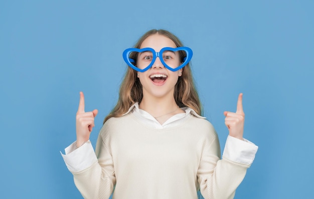 Fille heureuse dans de grands verres drôles en forme de coeur pointant les doigts vers le haut de l'espace de copie de fond bleu