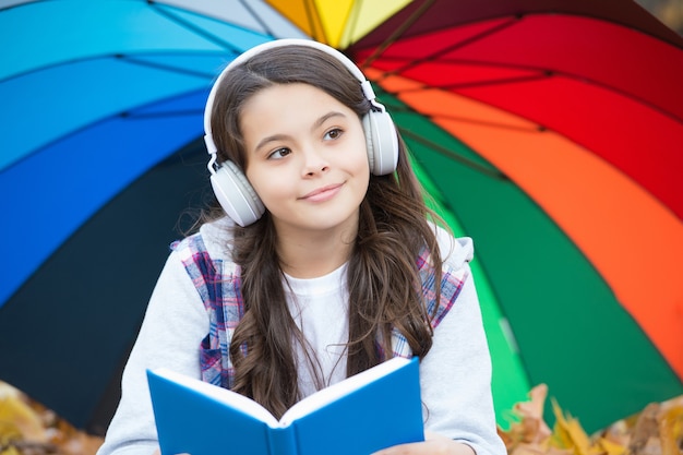 Une fille heureuse dans un style décontracté passe du temps à écouter de la musique et à lire un livre dans un parc d'automne, profitant du beau temps sous un parapluie coloré, de retour à l'école.