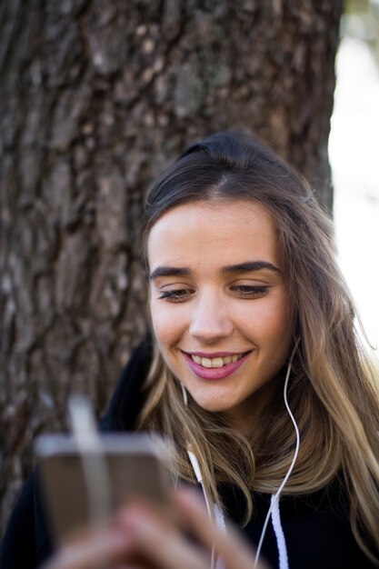 Fille heureuse, écouter de la musique en ligne avec des écouteurs sans fil d&#39;un smartphone contre l&#39;arbre