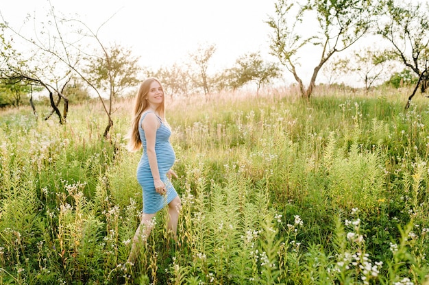 Fille heureuse enceinte aller, avec le ventre rond, aller sur l'herbe à l'extérieur dans la surface du jardin avec des arbres