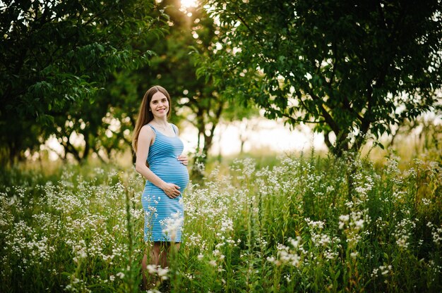 Fille heureuse enceinte se tenir debout et tenir la main sur le ventre, se tenir debout sur l'herbe à l'extérieur dans la surface du jardin avec des arbres