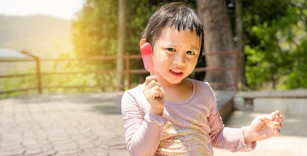 La fille heureuse d'enfant dans une chemise mouillée mangeant du popsicle de crème glacée dans le fond extérieur naturel.