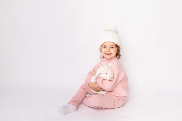 Fille heureuse d'enfant sur un fond d'isolement blanc avec un lapin de Noël dans ses mains dans un pull rose et un chapeau souriant, espace pour le texte