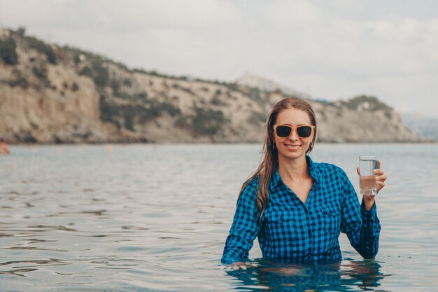 Une fille heureuse est assise dans la rivière et boit de l'eau dans un verre.