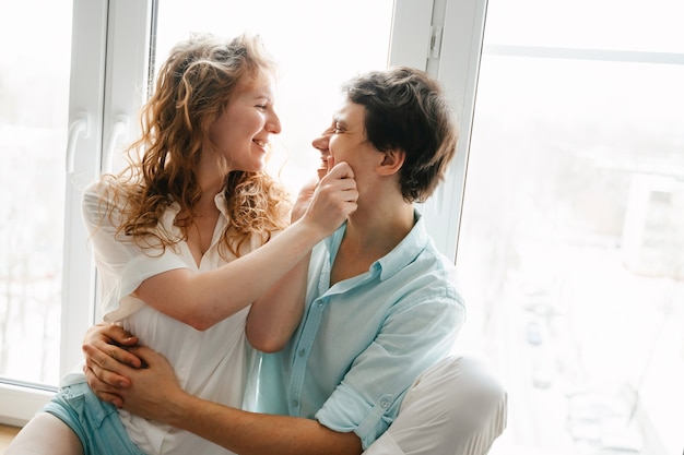 Fille heureuse et homme étreignant près de la fenêtre à la maison. Vêtements blancs et bleus. Saint Valentin.