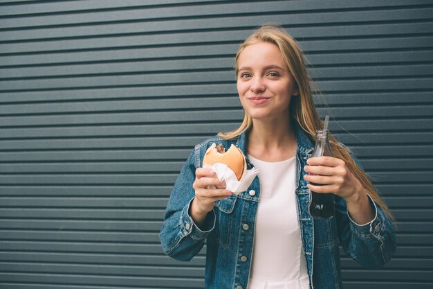 Fille heureuse mange de la restauration rapide et boit du cola près du mur gris