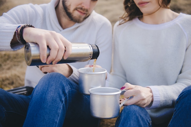 Fille heureuse et mec avec un sac à dos touristique et guitare assis dans un champ et boire du thé dans un thermos, concept d'histoire d'amour de voyage