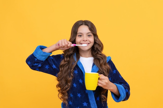 Une fille heureuse en peignoir confortable utilise une brosse à dents et une tasse, des soins personnels.