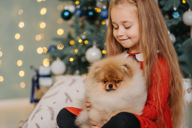 Fille heureuse posant avec un chiot mignon, arbre de Noël sur fond, cadeau moelleux tant attendu, joie