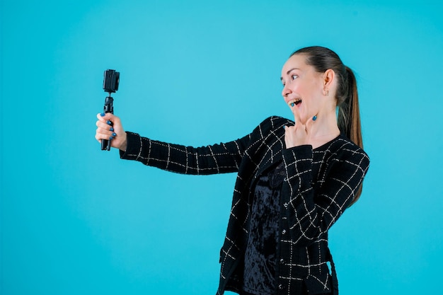 Une fille heureuse prend un selfie avec sa mini caméra en tenant l'index sur les lèvres sur fond bleu