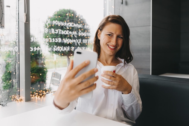Une fille heureuse prend un selfie sur un téléphone portable tout en mangeant un twister dans un café.