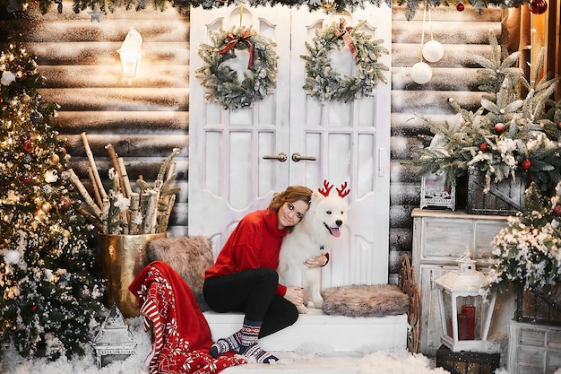 Fille heureuse en pull rouge de nouvel an et chaussettes scandinaves est assise sur les escaliers et posant avec mignon chien Samoyède blanc neige à l'intérieur de Noël. Concept de fête et décorations du nouvel an