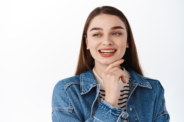 Fille heureuse réfléchie regardant loin l'espace de copie avec le texte promotionnel, souriant et touchant le menton tout en pensant, envisagez d'acheter le produit sur le lieu publicitaire de gauche, debout sur fond blanc