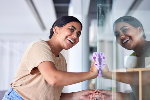 Fille heureuse et réflexion nettoyant la fenêtre avec un chiffon de ménage et un sourire satisfait pour une surface transparente brillante Concentration de bonheur et concentration de la jeune femme indienne polissant le verre dans la maison