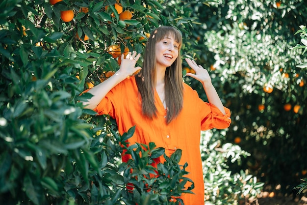Une fille heureuse en robe orange regarde la caméra en levant ses poignées dans un jardin d'orangers
