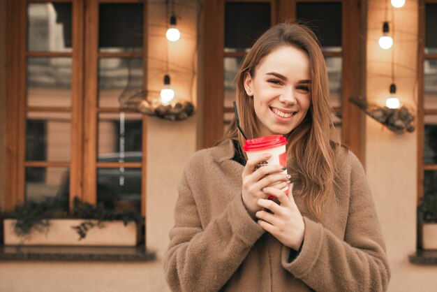 Fille heureuse se tient dans la rue avec une tasse de café dans ses mains, regarde la caméra, sourit, pose sur la caméra