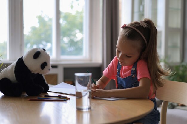 Une fille heureuse souriante assise à la table avec un ours panda jouet près d'elle, profitant d'une activité créative, dessinant des crayons à colorier des images dans des albums.