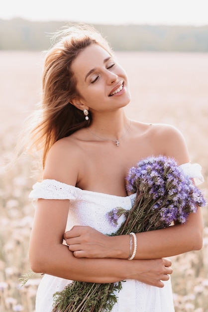 Fille heureuse sourit dans une robe blanche avec un bouquet de fleurs sur le terrain