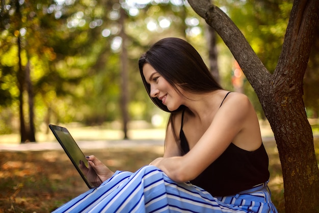 Fille heureuse avec une tablette se trouve sous un arbre, un grand portrait, regardez l'écran de la tablette