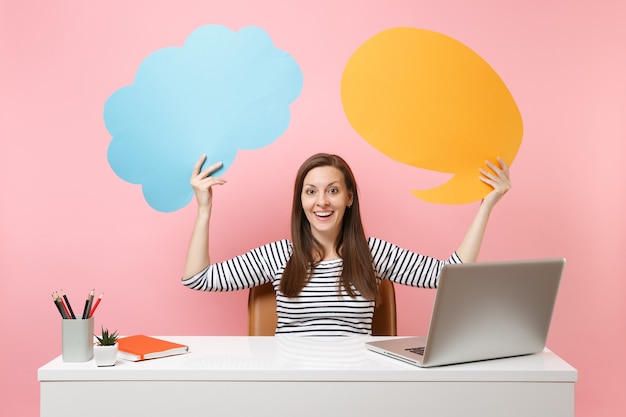 Une fille heureuse tient un blanc vide bleu jaune Dites que la bulle de dialogue nuage travaille au bureau blanc avec un ordinateur portable isolé sur fond rose pastel. Concept de carrière d'entreprise de réalisation. Copiez l'espace pour la publicité.