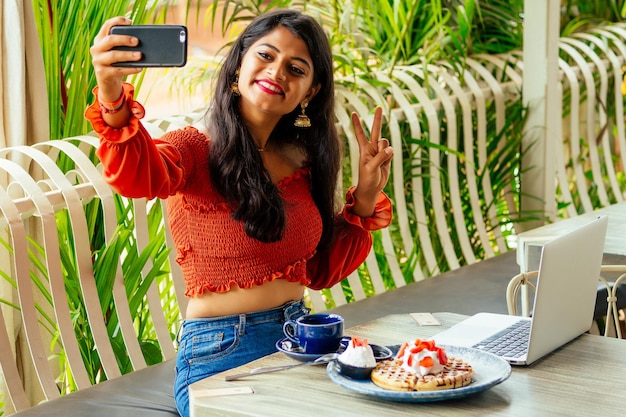 Fille hindoue en tenue moderne assise à une table de café avec un téléphone portable à la main faisant du selfie femme d'affaires mangeant des crêpes et travaillant sur un ordinateur portable à la véranda d'été l'étudiant de la pause déjeuner de l'université