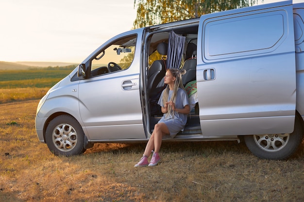 Une fille hippie est assise dans une mini-fourgonnette argentée sur un champ sur fond de montagnes le concept de f...