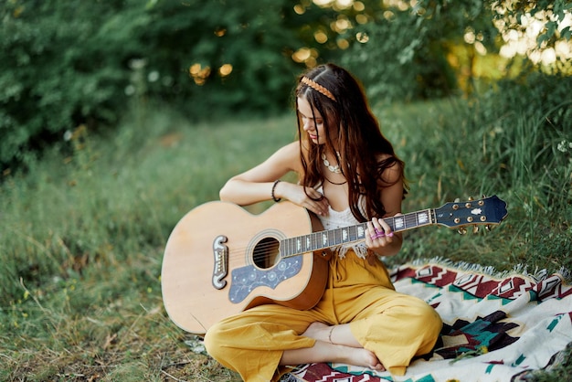 Fille hippie femme jouant de la guitare dans des vêtements écologiques assis sur le sol à l'extérieur dans la nature à l'automne en regardant le coucher du soleil