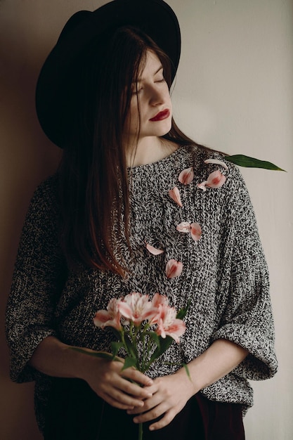 Fille hipster élégante au chapeau tenant des fleurs roses dans la chambre boho femme tenant belle alstroemeria dans les mains au printemps matin et pétales sur le corps portrait féminin sensuel créatif