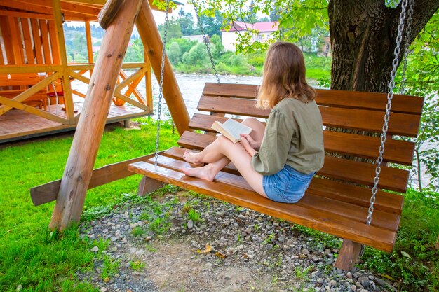 Fille hipster lit un livre sur un banc en bois. Femme détente à l'extérieur sur la balançoire du porche dans la nature. Photo confortable. Ambiance estivale.