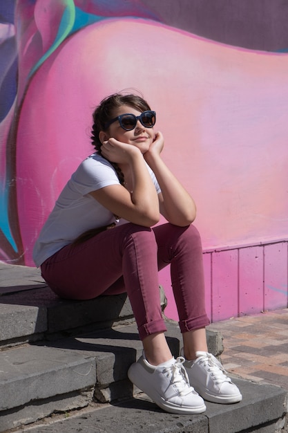 Photo fille de hipster sur des lunettes de soleil assis sur des marches sur fond rose