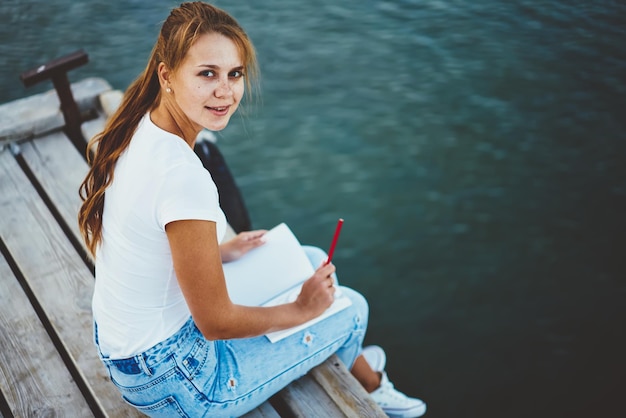 fille hipster positive écrivant un essai avec une idée dans un cahier alors qu'elle était assise sur une jetée en bois près du lac