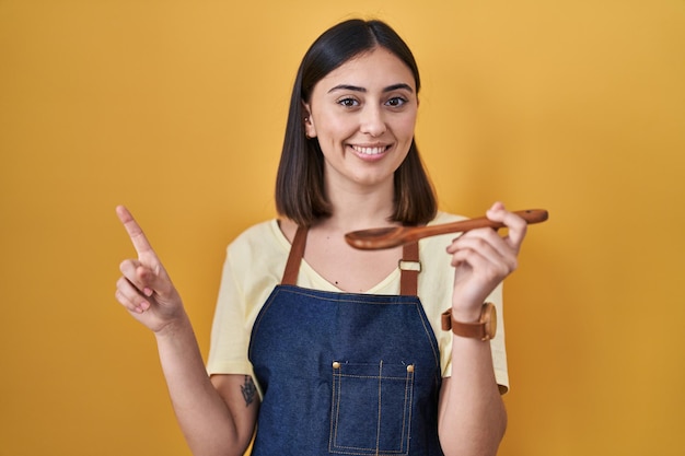 Fille hispanique mangeant une cuillère en bois saine avec un grand sourire sur le visage, pointant avec le doigt de la main sur le côté en regardant la caméra.