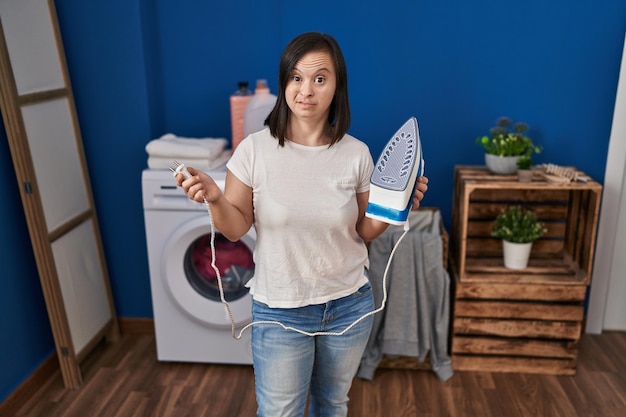 Fille hispanique avec le syndrome du duvet repassant des vêtements à la maison en souriant regardant sur le côté et en regardant loin en pensant.