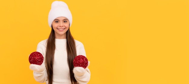 Fille d'hiver heureux enfant en chapeau d'hiver et gants tenir des boules décoratives du nouvel an sur fond jaune Noël Bannière de noël enfant fille studio enfant hiver portrait avec espace de copie